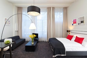 Et hotelværelse med en seng, en sofa, et sofabord med blomster, et skrivebord med en skål frugt og et stort vindue med gardiner.