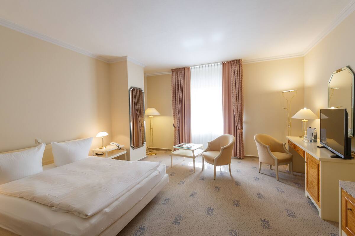 A hotel room with a large bed, two chairs, a desk with a mirror, and a television.