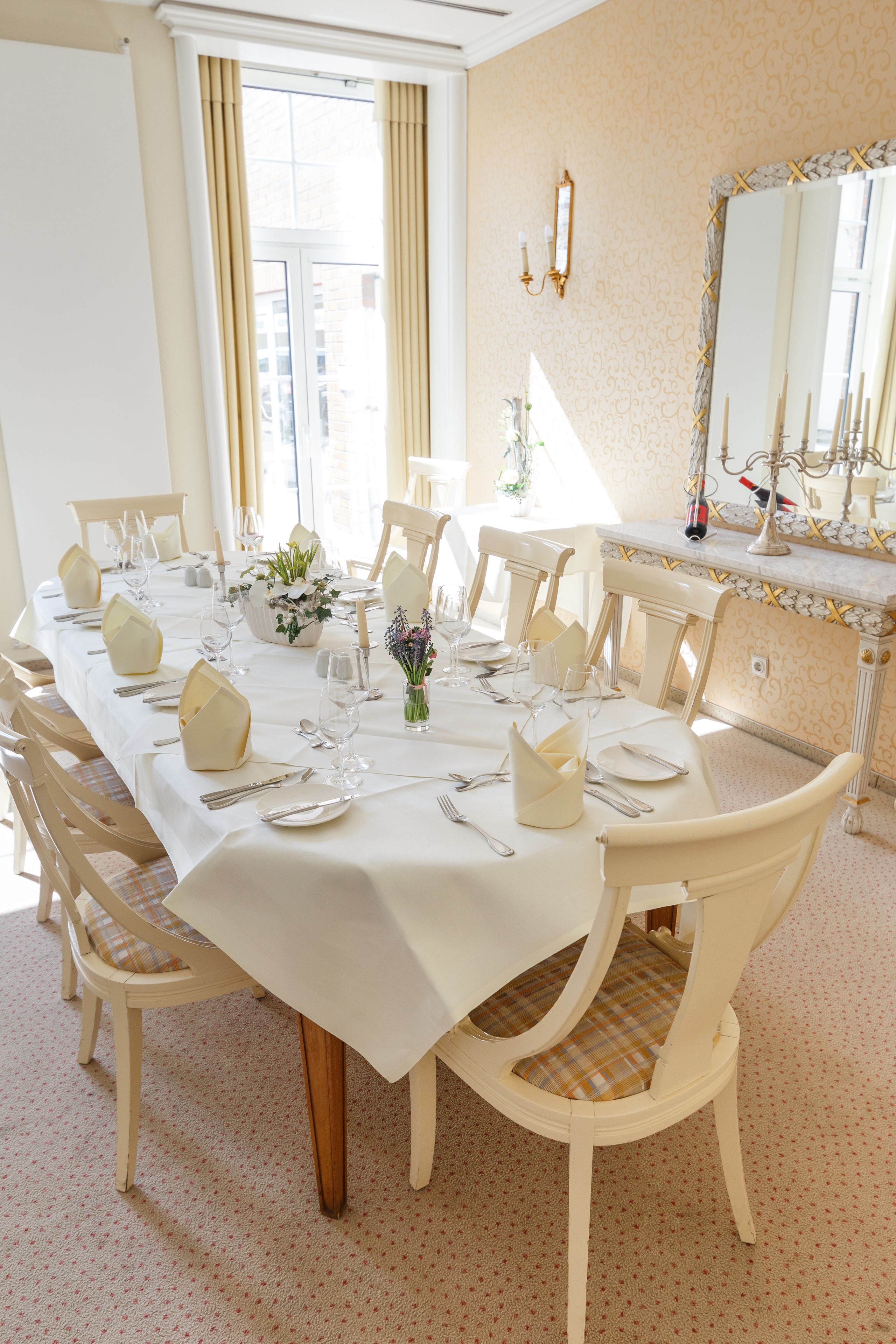 A dining room with a long table set for a meal, chairs, a vase with flowers, and a mirror on the wall.