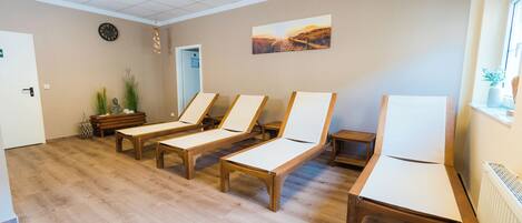 A room with wooden flooring, four wooden lounge chairs with white cushions, a clock on the wall, and a painting above a window.
