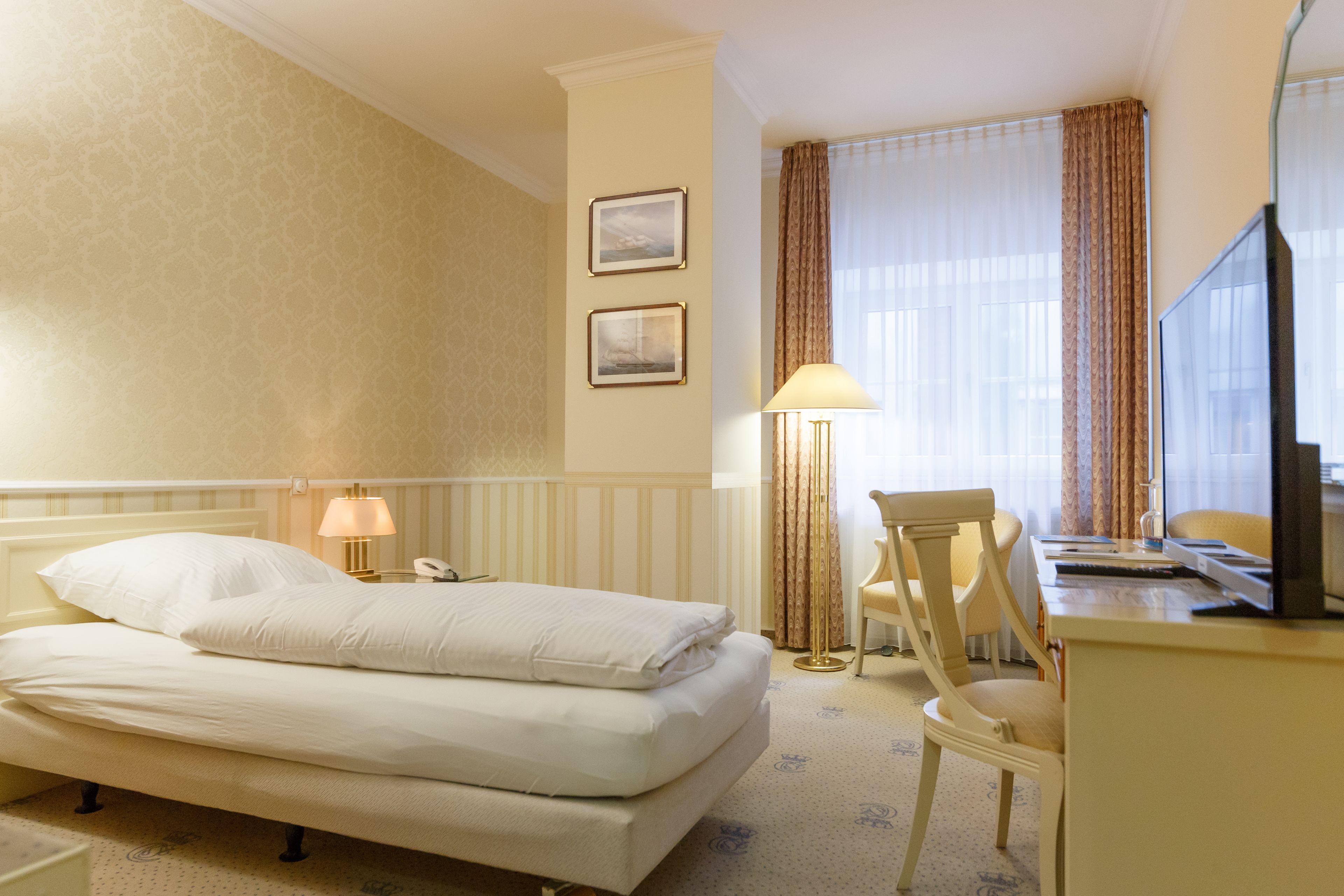 A hotel room with a bed, a desk, a chair, a lamp, and two framed pictures on the wall.