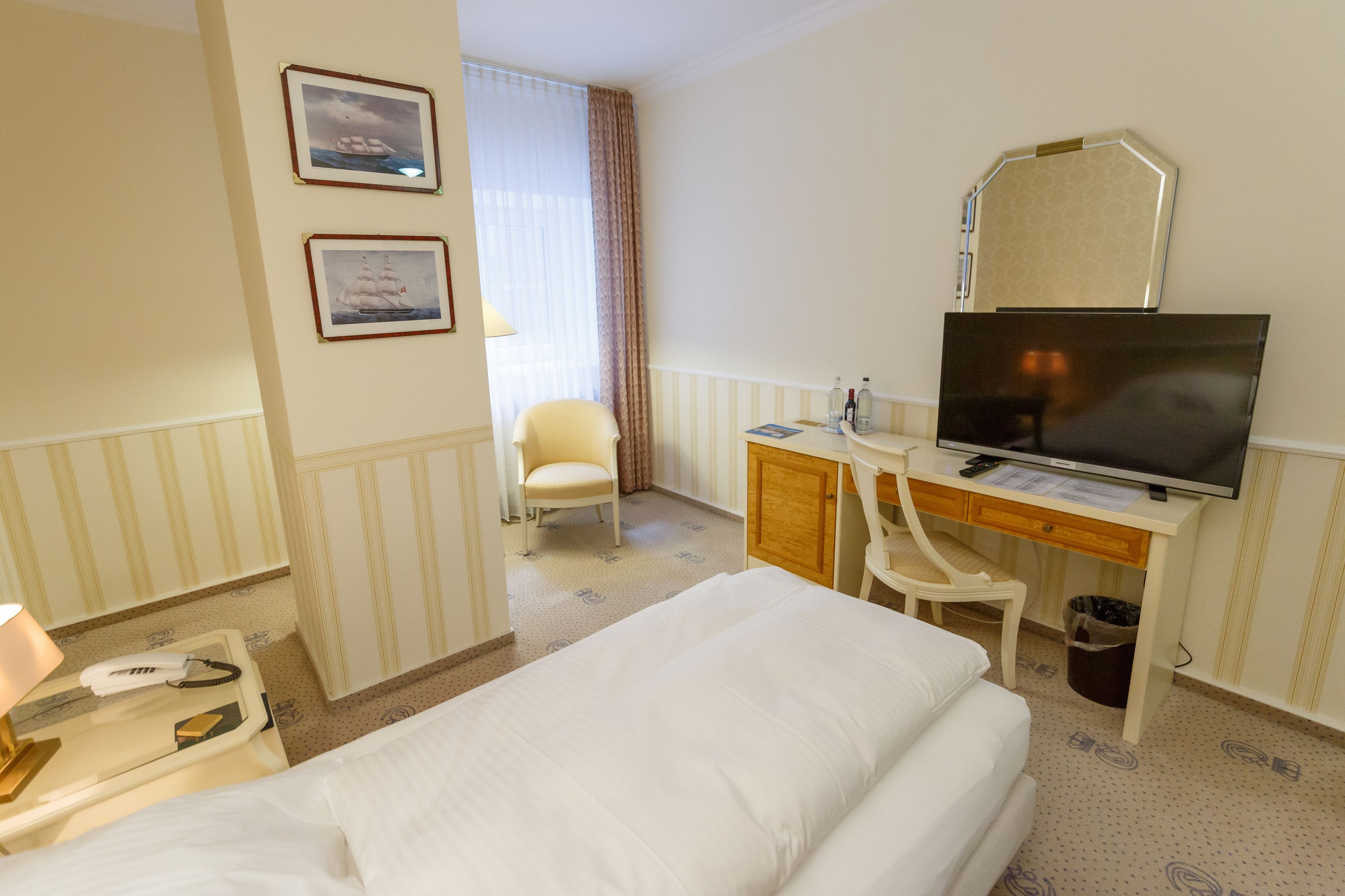 A hotel room with a bed, a desk with a computer, a chair, a desk lamp, a telephone, and a trash can.