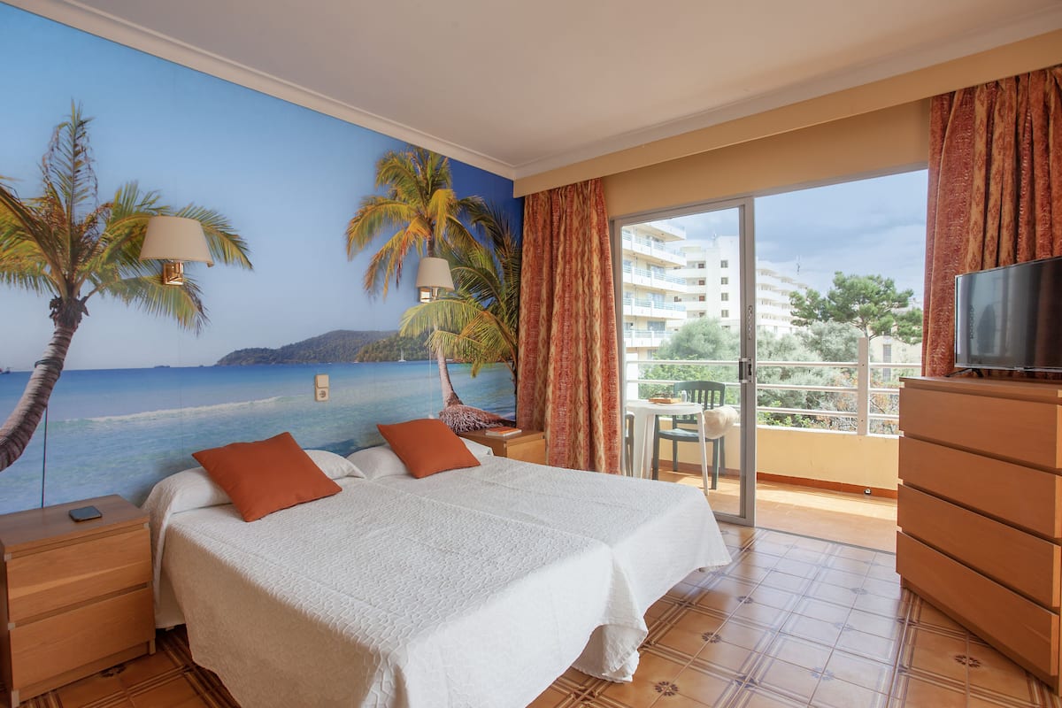 A hotel room with a bed, a TV, a nightstand, a dresser, and a balcony with a view of the ocean and buildings.