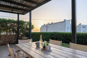 An outdoor dining area with a wooden table, chairs, a bottle, glasses, and a bowl of fruit.