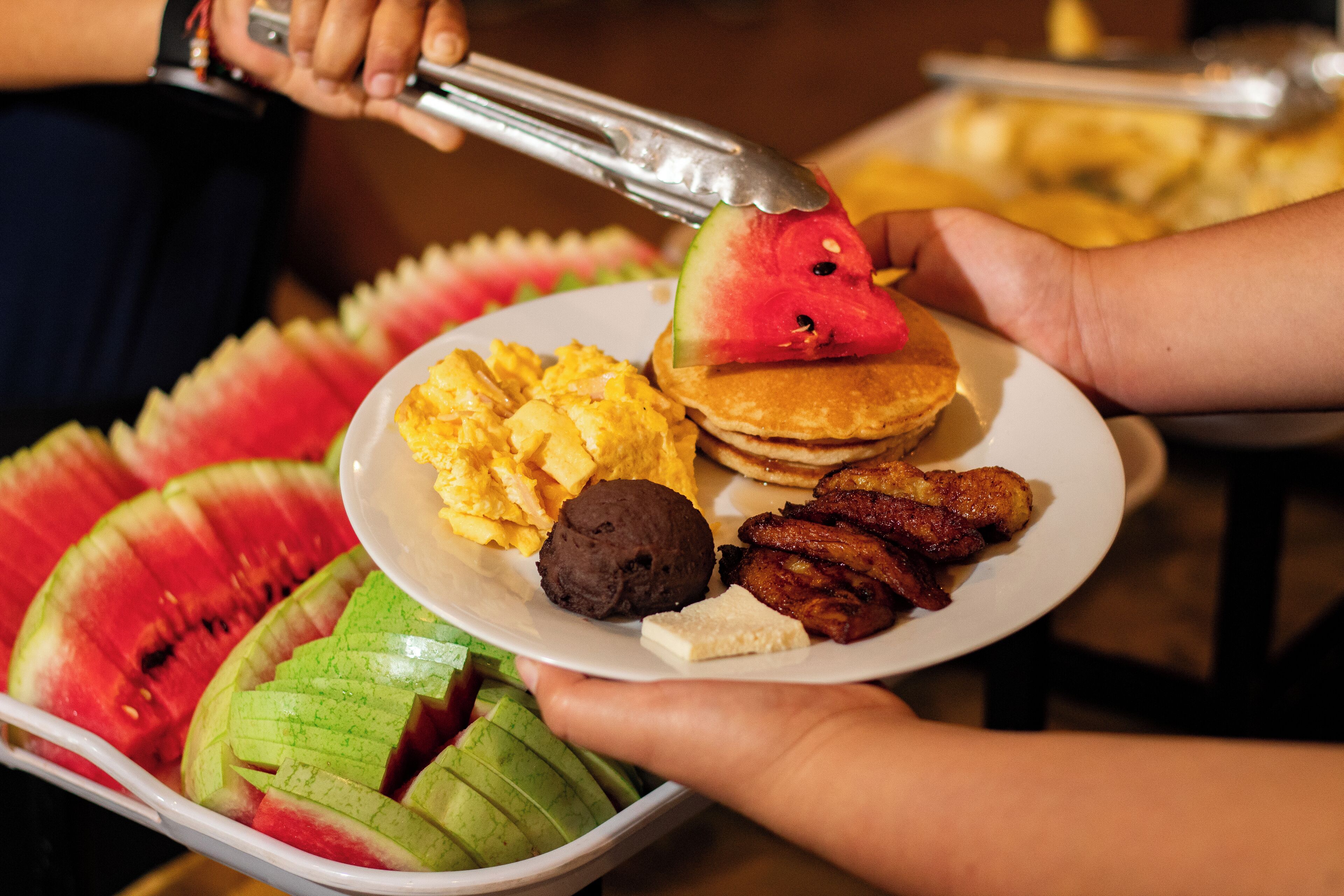 Un plato con huevos revueltos, panqueques, tocino, una porción de helado y una rebanada de sandía.