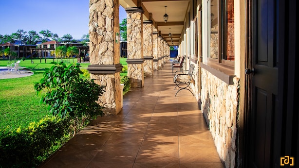 A covered outdoor corridor with stone pillars, chairs, and a view of a landscaped area. - Grand Caporal Hotel (Chiquimula)
