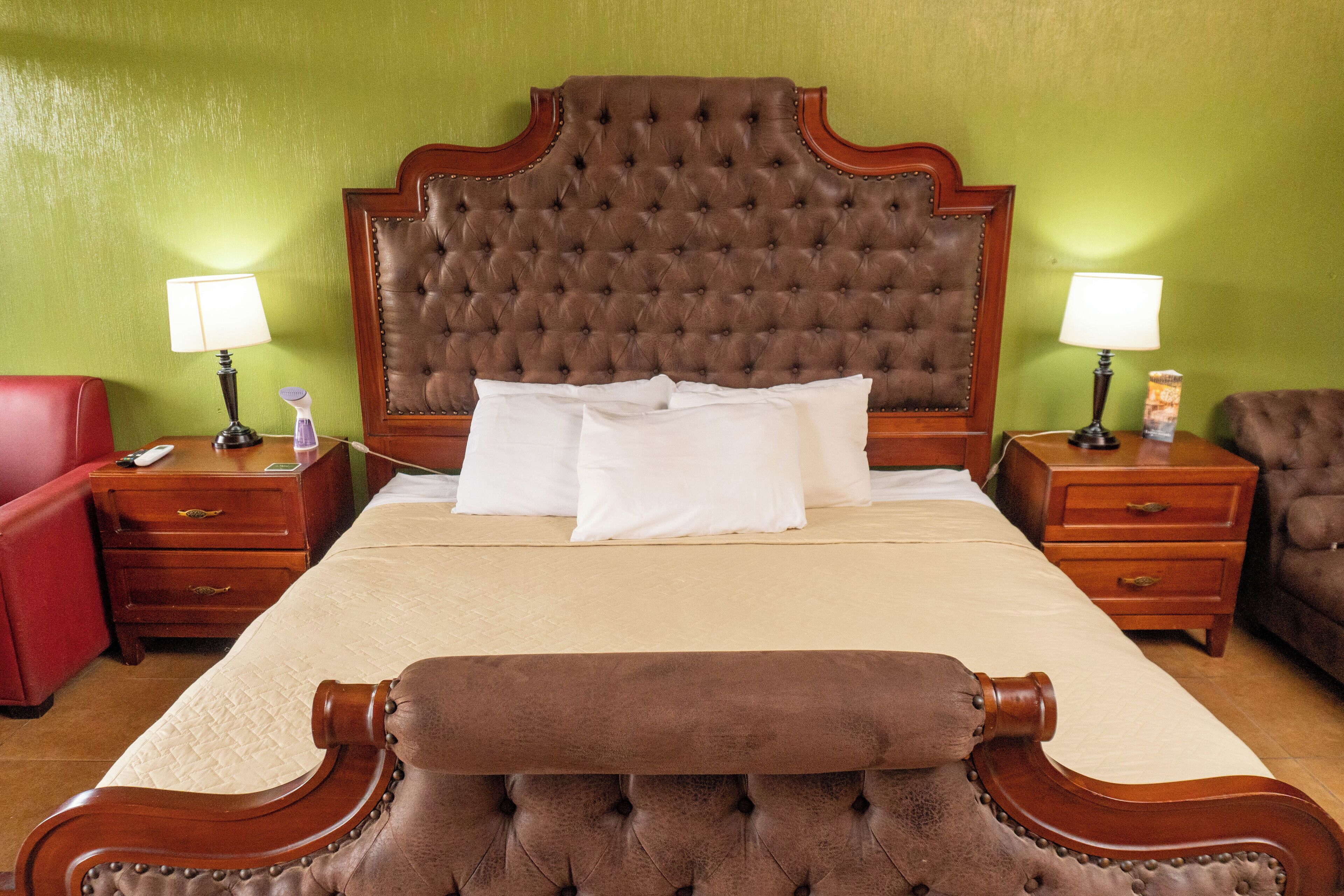 A hotel room with a large tufted headboard, a wooden bench, a red armchair, and two bedside tables with lamps and a small decorative item.