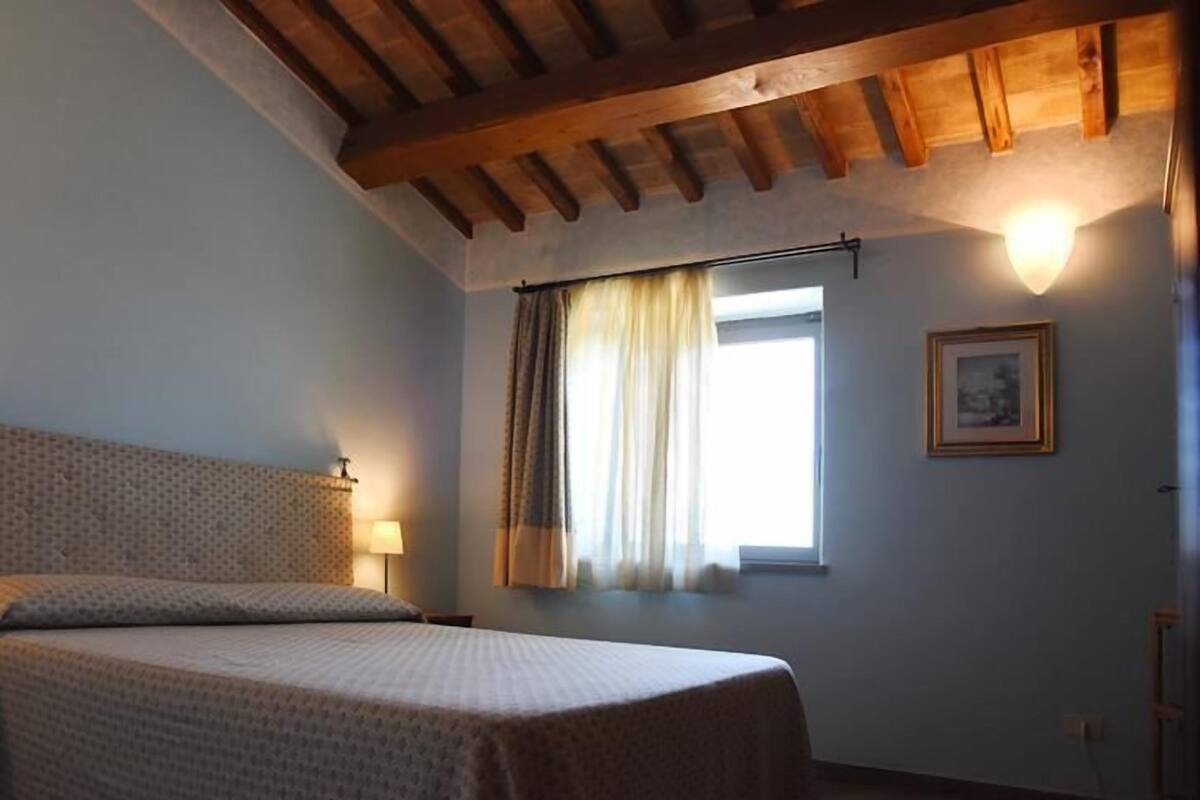 A bedroom with a bed, a window with curtains, a wall-mounted light, and a framed picture.