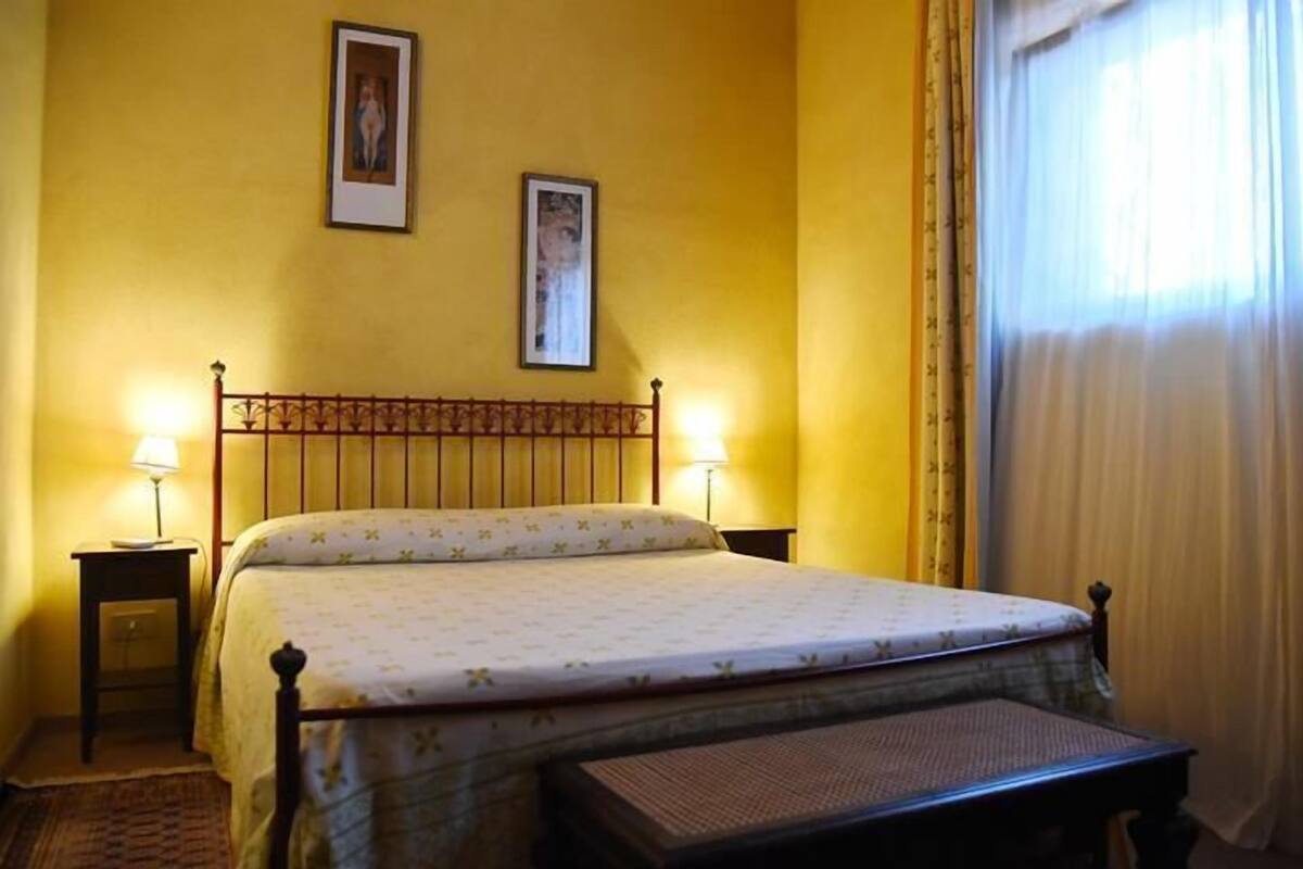 A bedroom with a bed, two bedside tables, a nightstand, a bench, and two framed pictures on the wall.