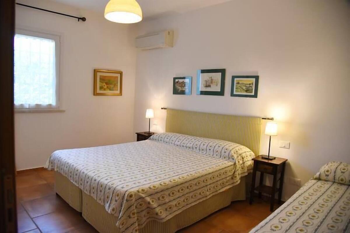 A bedroom with a large bed, two bedside tables with lamps, a window with curtains, and three framed pictures on the wall.