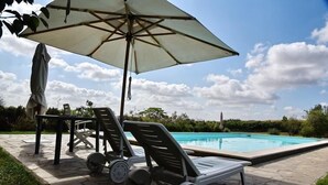 Un área junto a la piscina con sillones de descanso y una sombrilla, rodeada por un patio de piedra y vegetación.