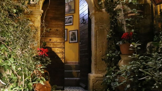 Un arco de piedra que da a una puerta de madera, rodeado de macetas con plantas y un cartel con un nombre.
