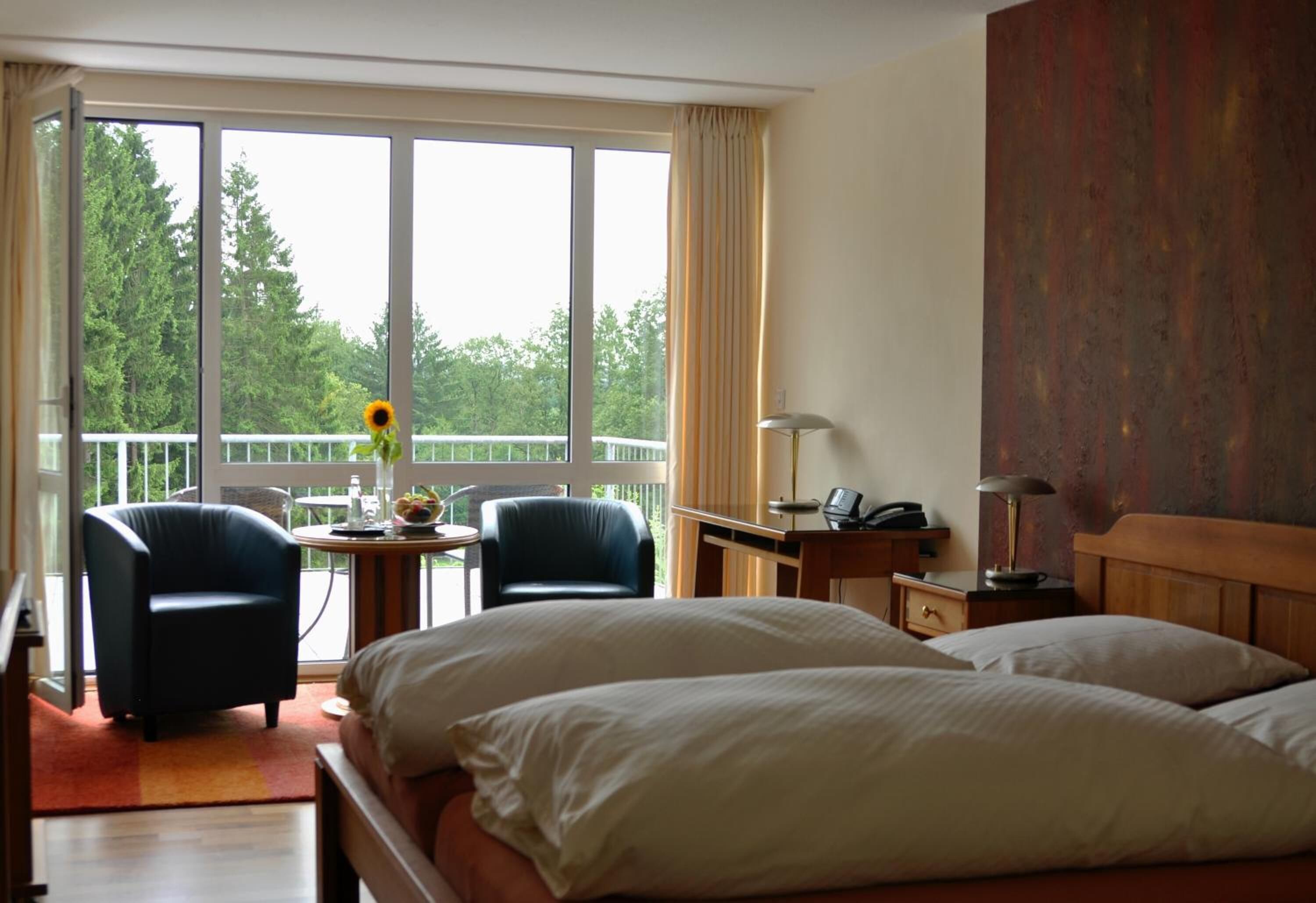 A hotel room with a large window, a bed, two armchairs, a small table with a vase, and a desk with a lamp.