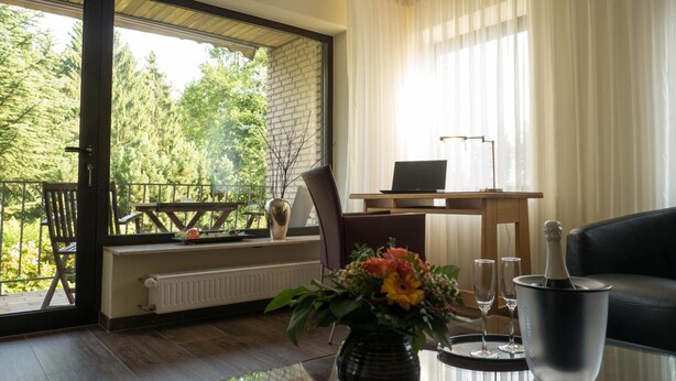 Una sala de estar moderna con una mesa de centro de vidrio, un ordenador portátil sobre un escritorio de madera, un sofá de cuero negro, y vistas a un balcón con una mesa y sillas de madera.
