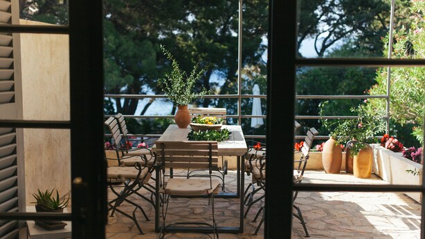 An outdoor patio with a stone floor, a wooden table, and metal chairs. There are potted plants and a view of trees through large windows. - Seafront Villa in Bol - Prime Location  (Bol, island of Brac)