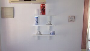 Een kamer met een tafel en twee stoelen, een plank met decoratieve objecten, een airconditioner en een raam met gordijnen.