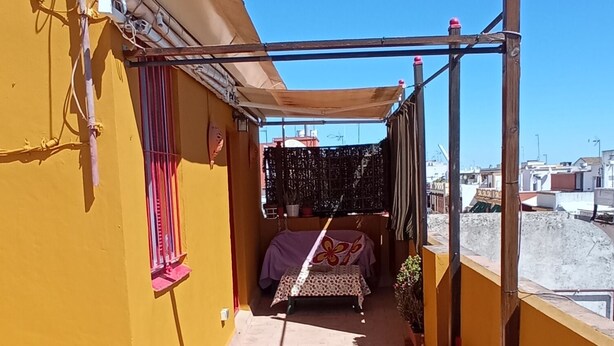 Een terras op het dak met een gele muur, een hangstoel en een tafel met een tafelkleed met patroon.