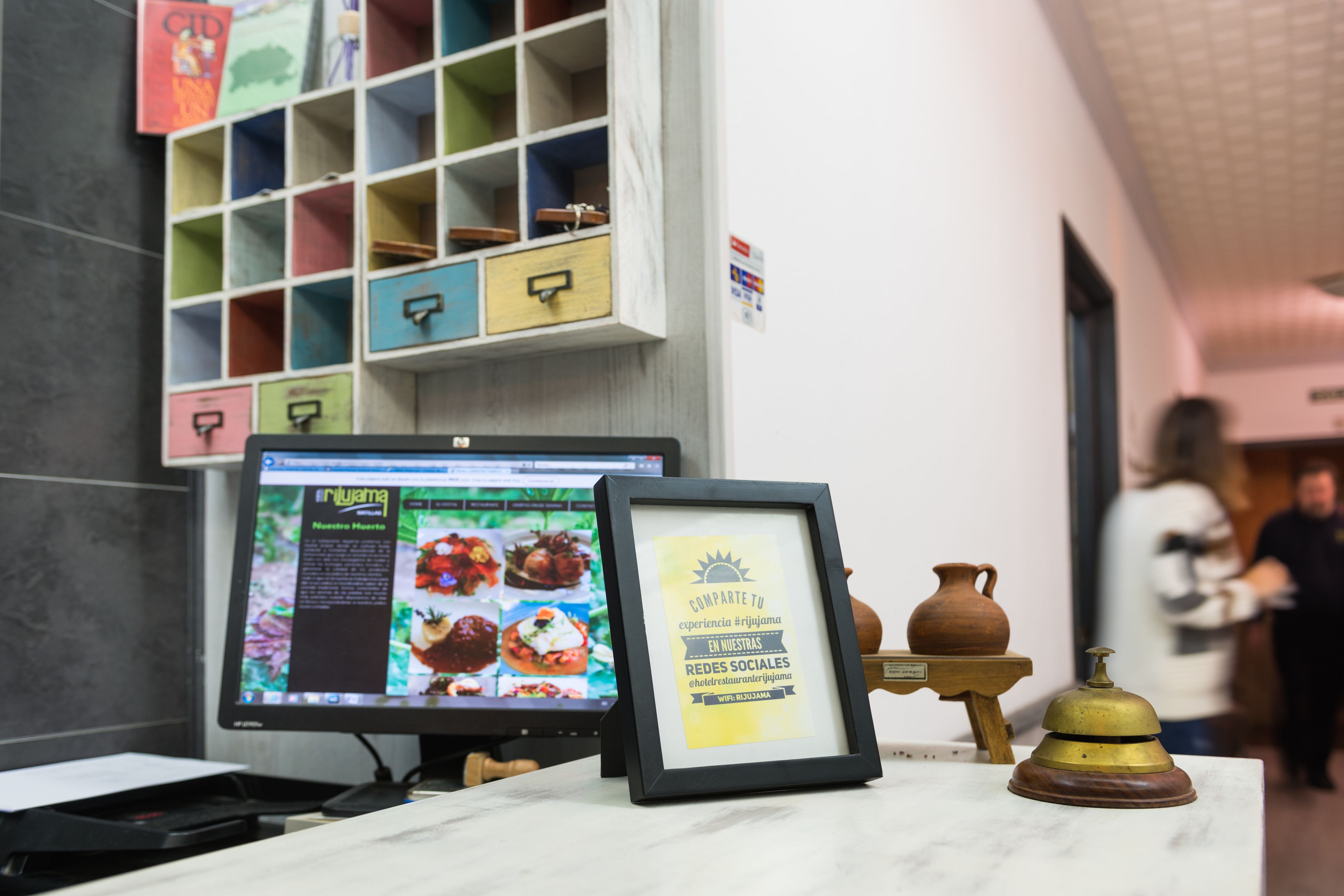 Un mostrador de recepción con un monitor de computadora que muestra un sitio web, un cartel enmarcado y una pequeña campana sobre un soporte.