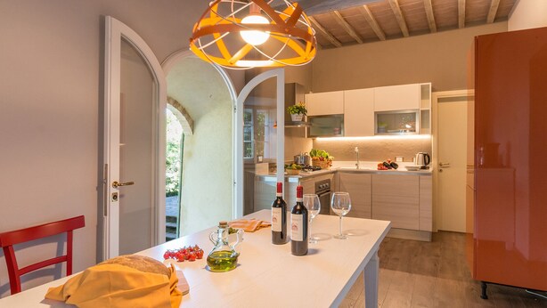 Uma cozinha moderna com armários brancos, uma mesa de jantar com toalha amarela e uma cadeira vermelha. Há garrafas de vinho e copos sobre a mesa.