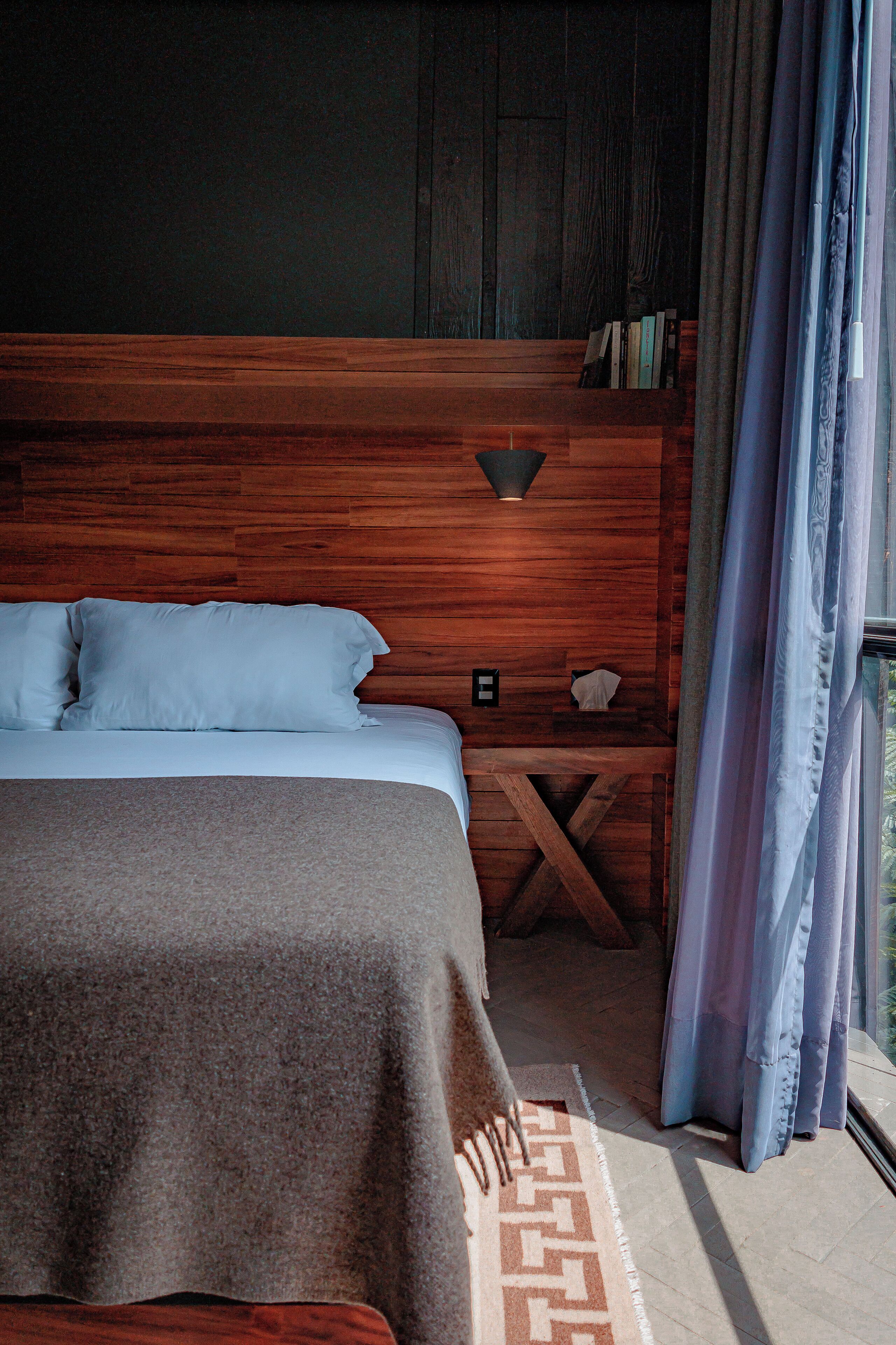 A bedroom with a wooden headboard, a bed with white linens, a nightstand with a lamp, and a view through a window with curtains.