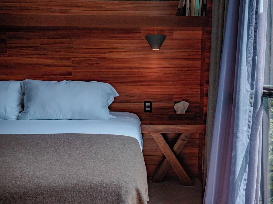 A bedroom with a wooden headboard, a bed with white linens, a nightstand with a lamp, and a view through a window with curtains.