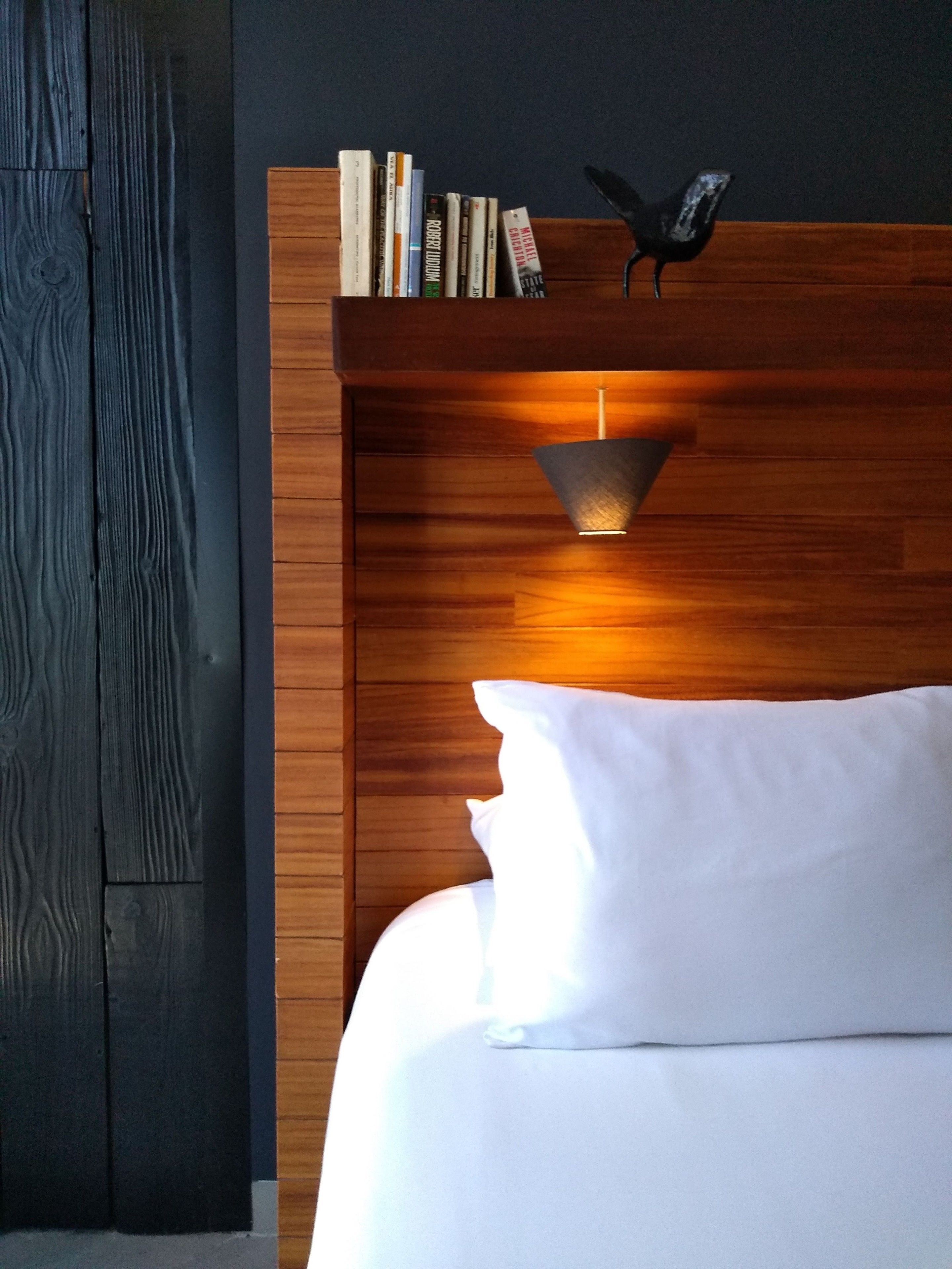 Ein ordentlich bezogenes Bett mit weißen Leinen, einem Holz-Kopfteil und einem Regal mit Büchern und einem dekorativen Vogel.