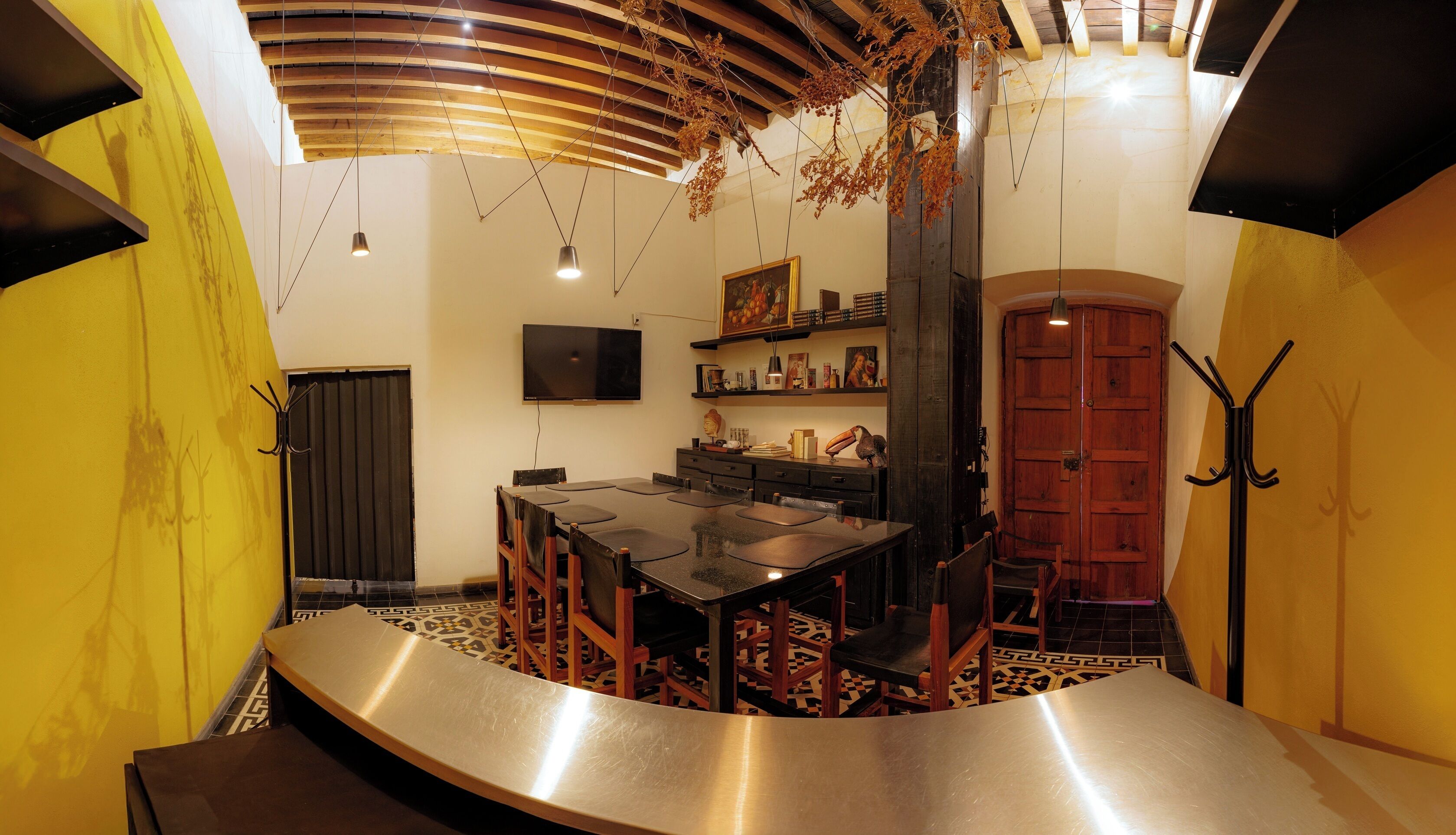 A dining area with a curved counter, wooden chairs, and a black table. There is a television mounted on the wall and a wooden door in the background.