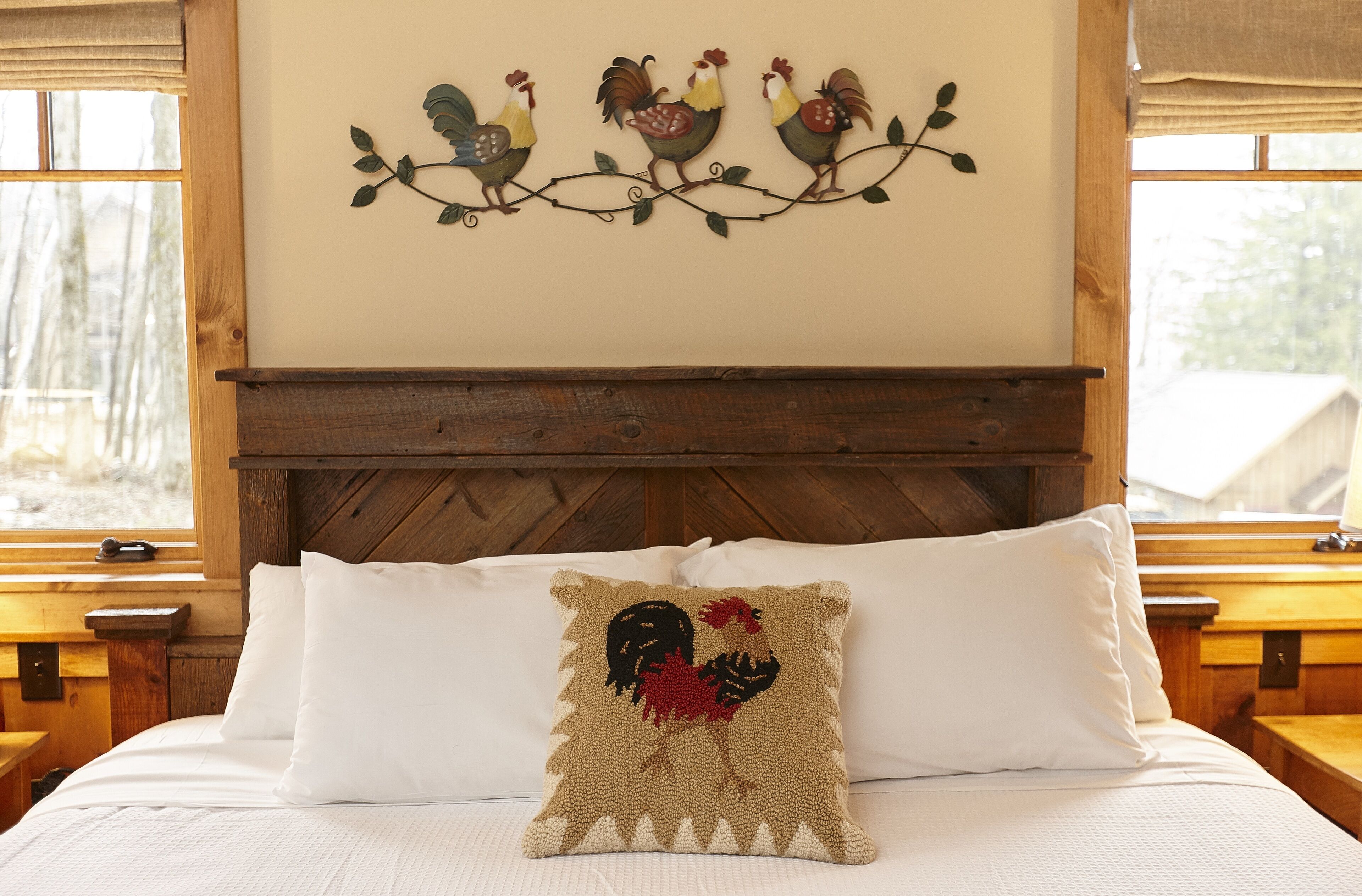 A bed with white linens, a wooden headboard, and a decorative pillow with a rooster design.