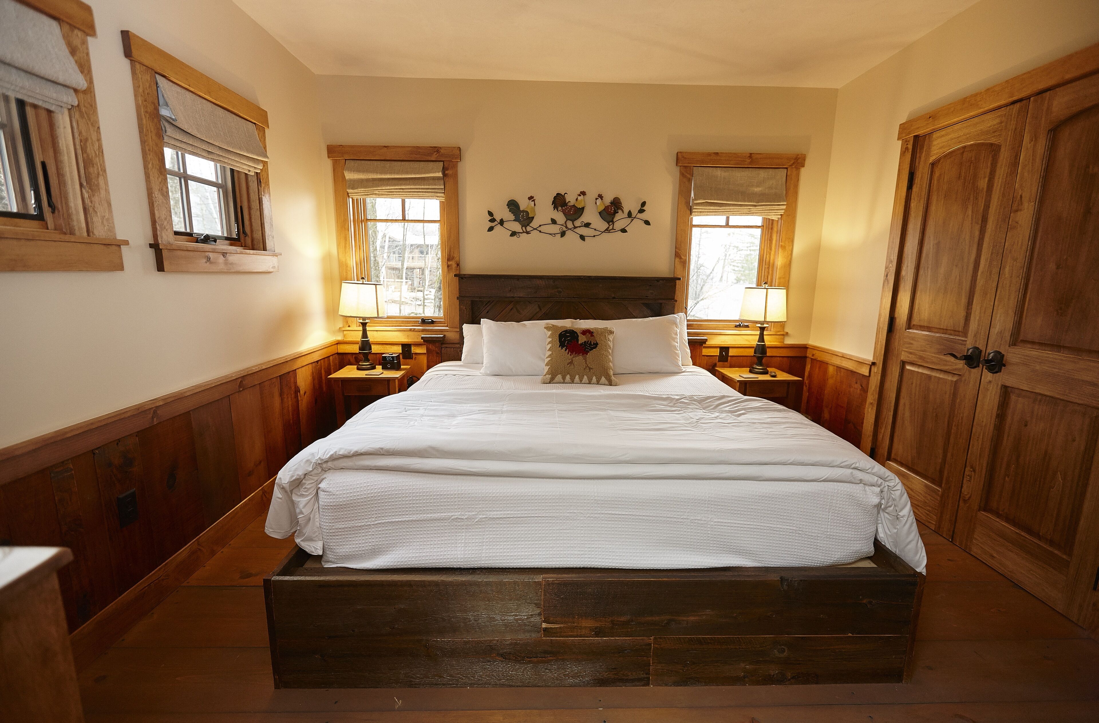 A large wooden bed with a white bedspread and a decorative pillow, flanked by wooden nightstands with lamps, in a room with wooden walls and a wooden door.