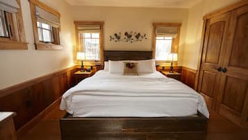Een groot houten bed met een witte spreidek en een decoratief kussen, aan weerszijden met houten nachtkastjes met lampen, in een kamer met houten wanden en een houten deur.