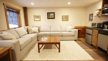 A living room with a sectional sofa, a wooden coffee table, and a kitchen area with appliances.