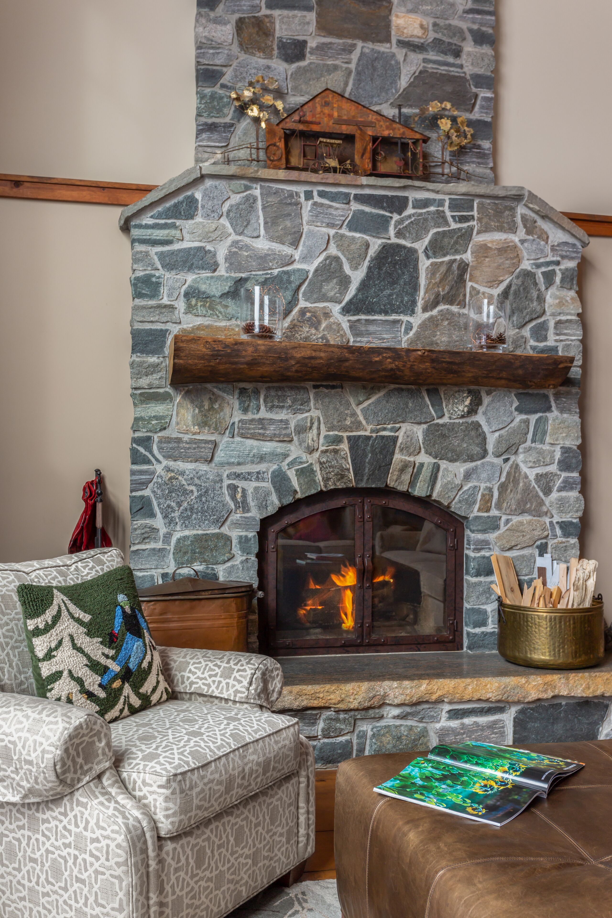 Una chimenea de piedra con repisa de madera, un rincón de estar acogedor con cojines estampados y un adorno sobre la chimenea.