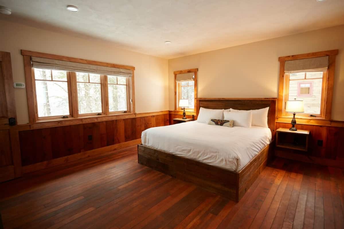 A bedroom with a wooden bed frame, a large window, and a nightstand with a lamp.