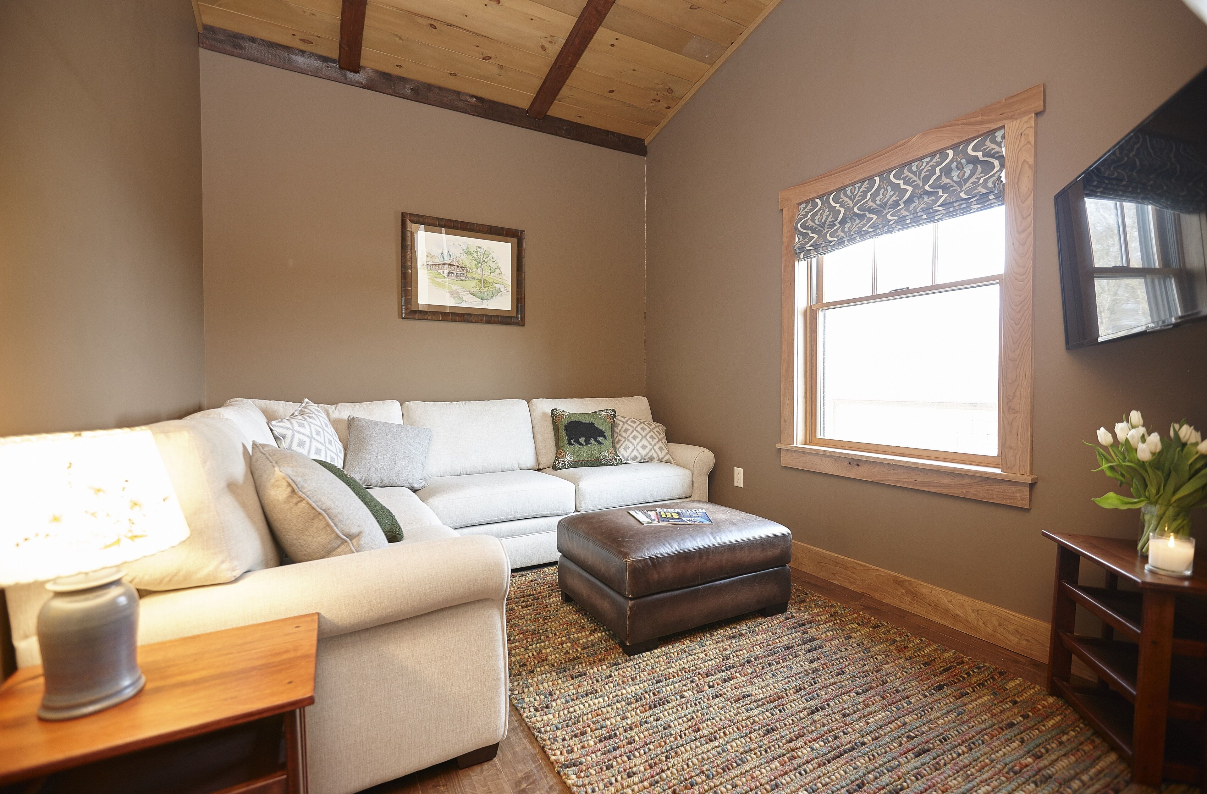 A cozy living room with a beige sofa, a brown ottoman, a patterned rug, a wooden floor, and a window with a decorative valance.
