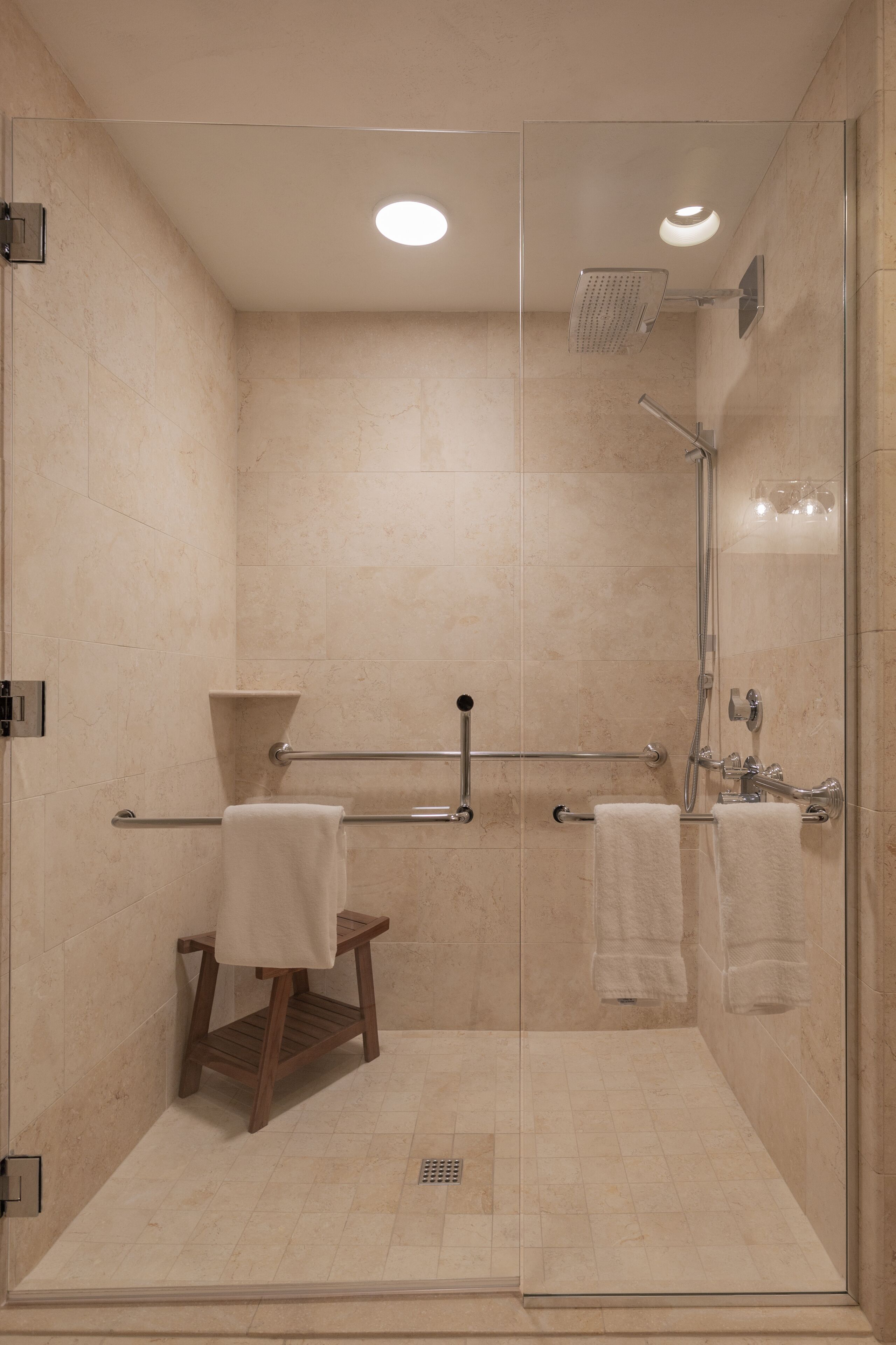 A walk-in shower with a glass door, a wooden stool, and two towels hanging on a towel bar.