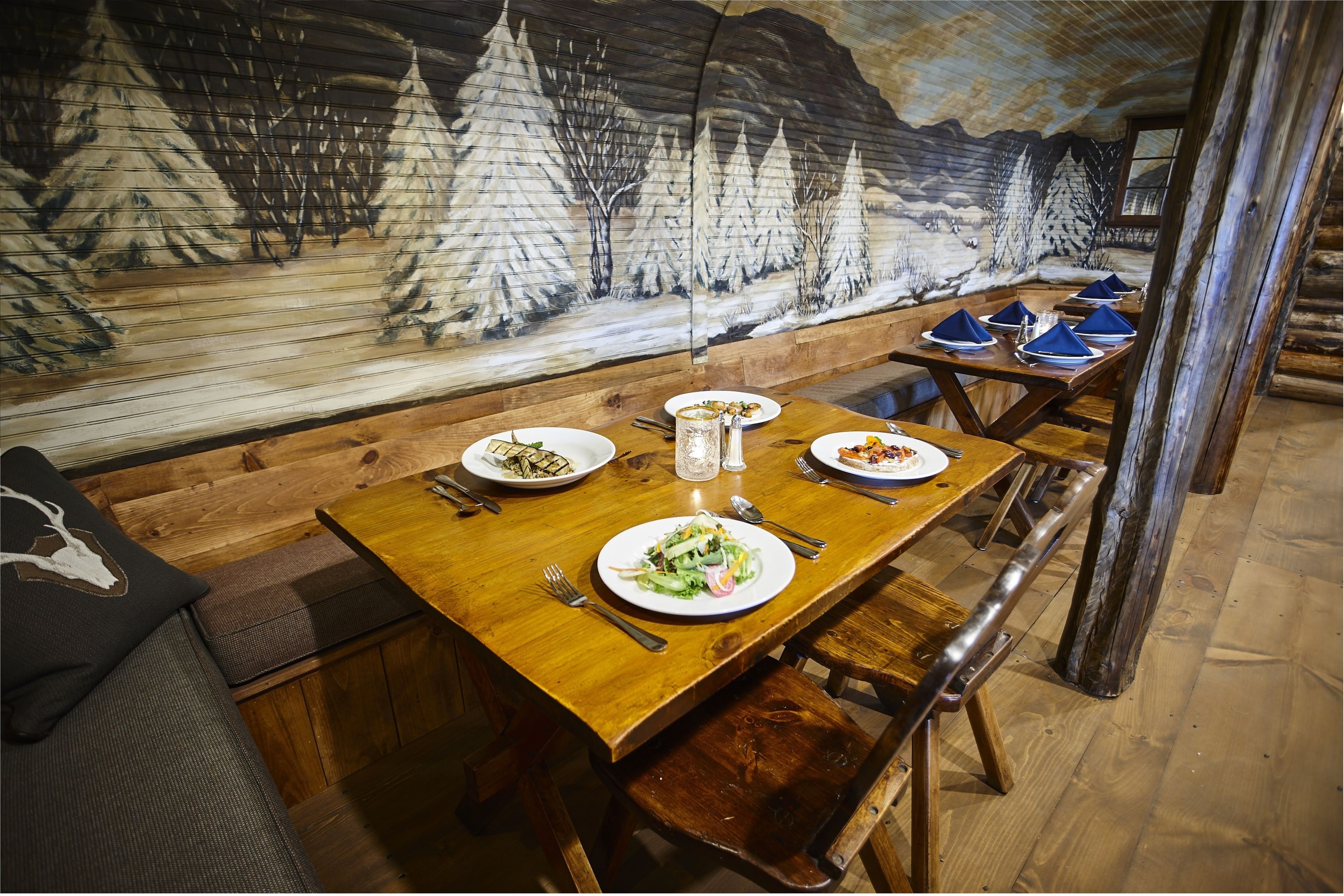 A wooden dining table with plates of food and cutlery, set against a mural of a snowy landscape.
