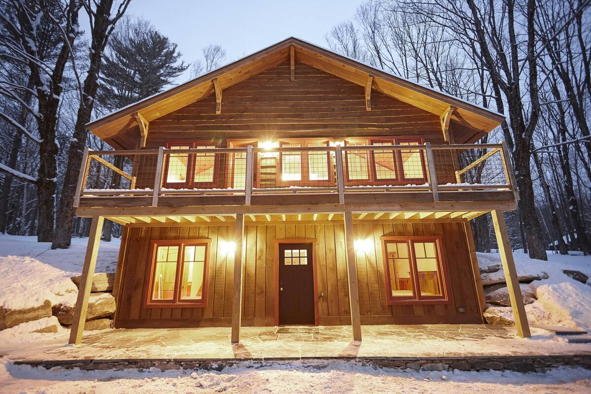 A two-story wooden cabin with a balcony, surrounded by snow and trees.