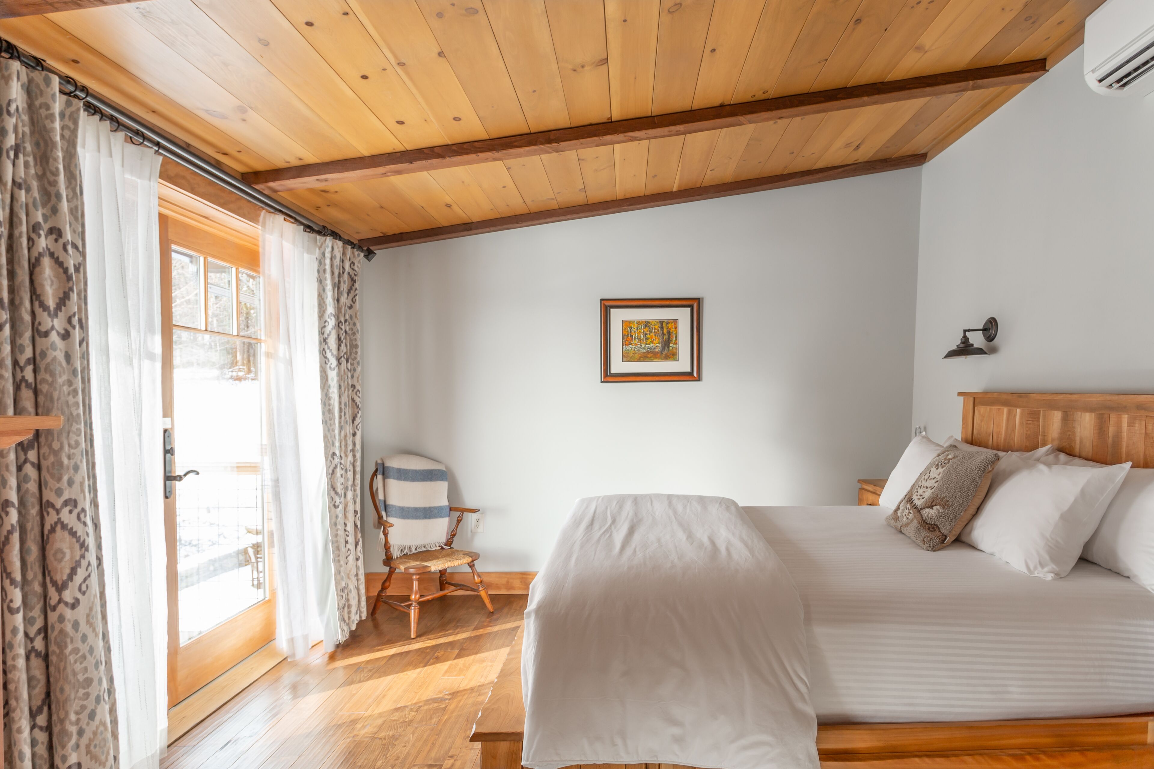 A bedroom with a wooden ceiling, a bed with white bedding, a chair, a framed picture on the wall, and a window with curtains.