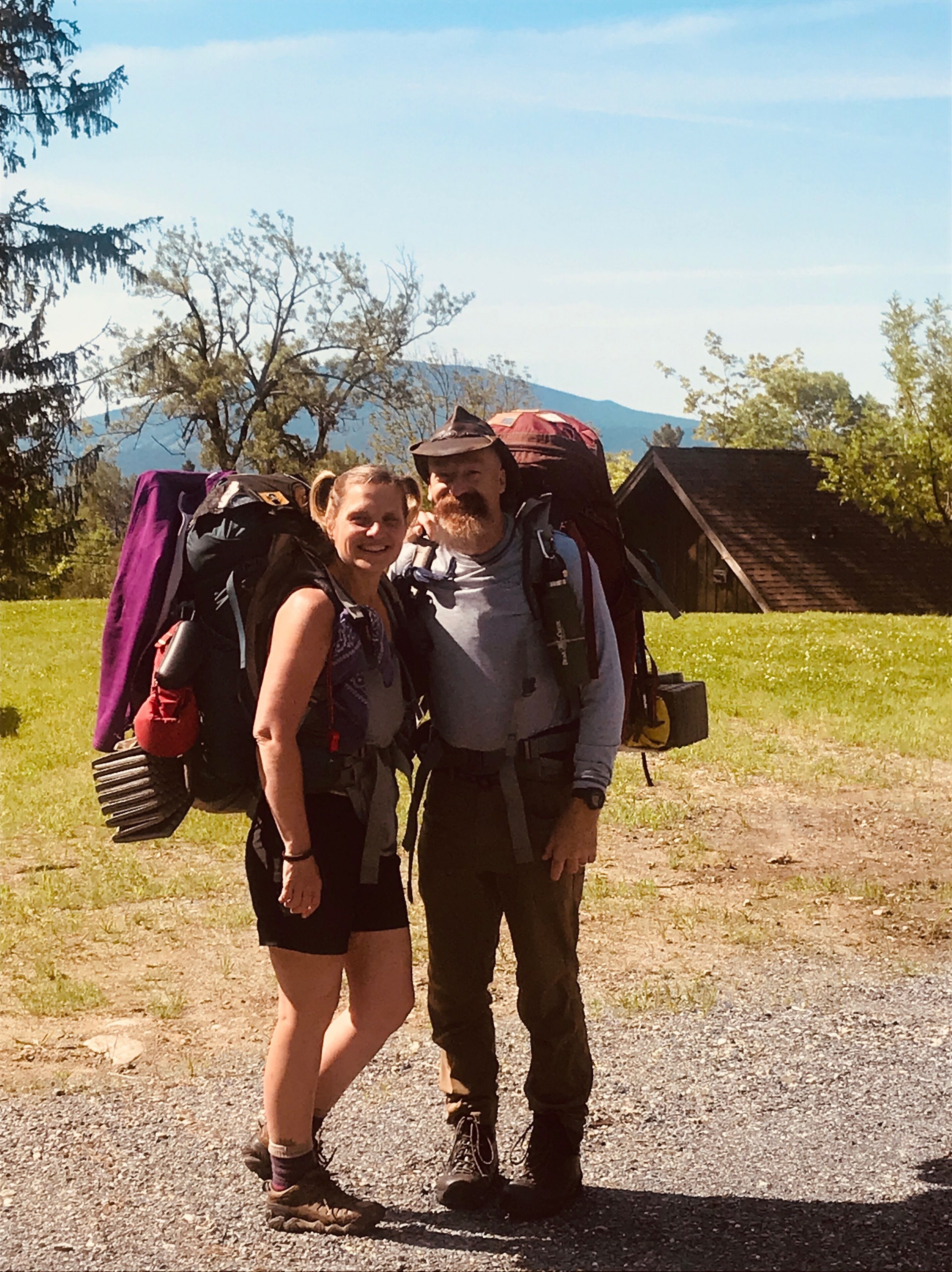 Two people with backpacks standing on a gravel path with a wooden structure and trees in the background.