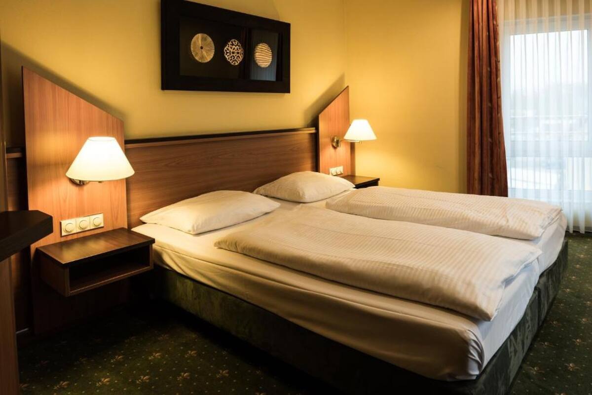 A hotel room with two beds, a wooden headboard, bedside lamps, a small shelf, and a wall-mounted clock.