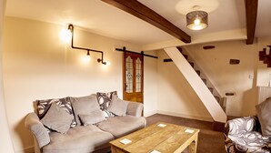Un salon chaleureux avec un canapé, une table basse en bois et un fauteuil à motifs. On aperçoit un escalier au fond de la pièce.