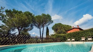 Une piscine entourée d’une clôture en bois, avec des chaises longues et, au fond, un bâtiment doté d’un toit rouge.
