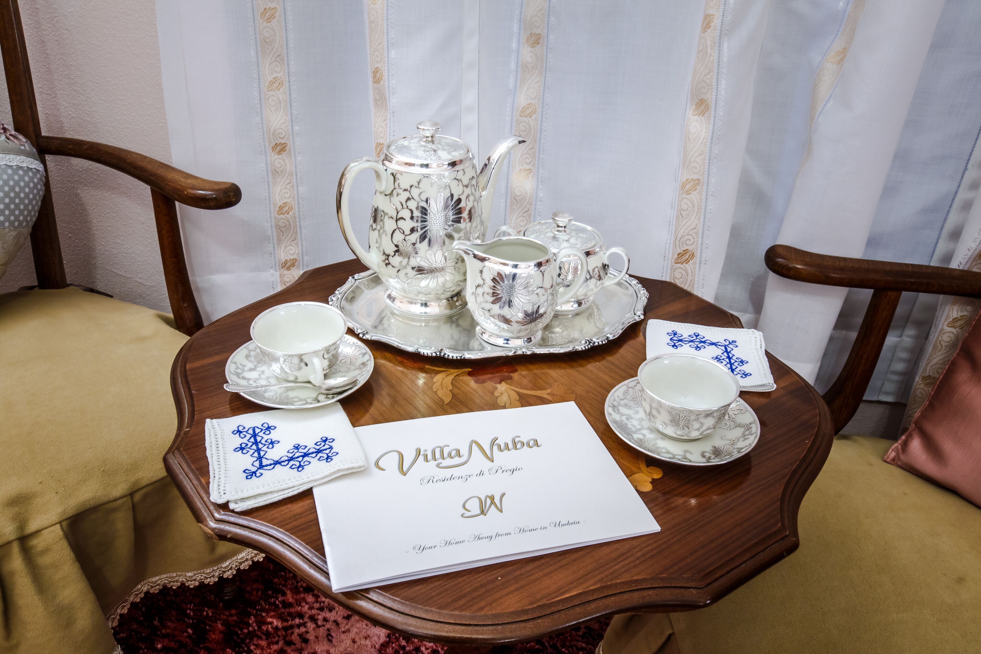 A table set with silver tea service, a white napkin with blue embroidery, and a sign reading 'Villa Nuba Residence di Pregio.'