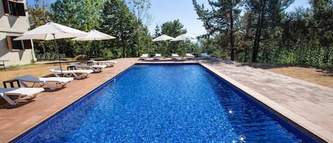 Piscina rectangular con agua azul cristalina, rodeada de sillas de descanso y sombrillas, con árboles y un edificio de fondo.