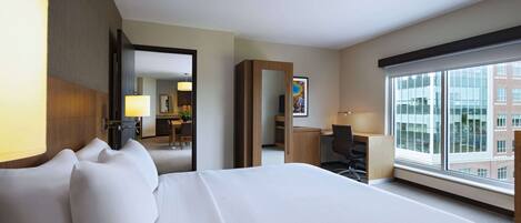 A hotel room with a large bed, a desk, a chair, a desk lamp, a large window with a city view, and a mirror.