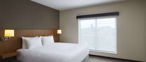 A hotel room with a large bed, two bedside lamps, a window with a view of a cityscape, and a dark-colored headboard.