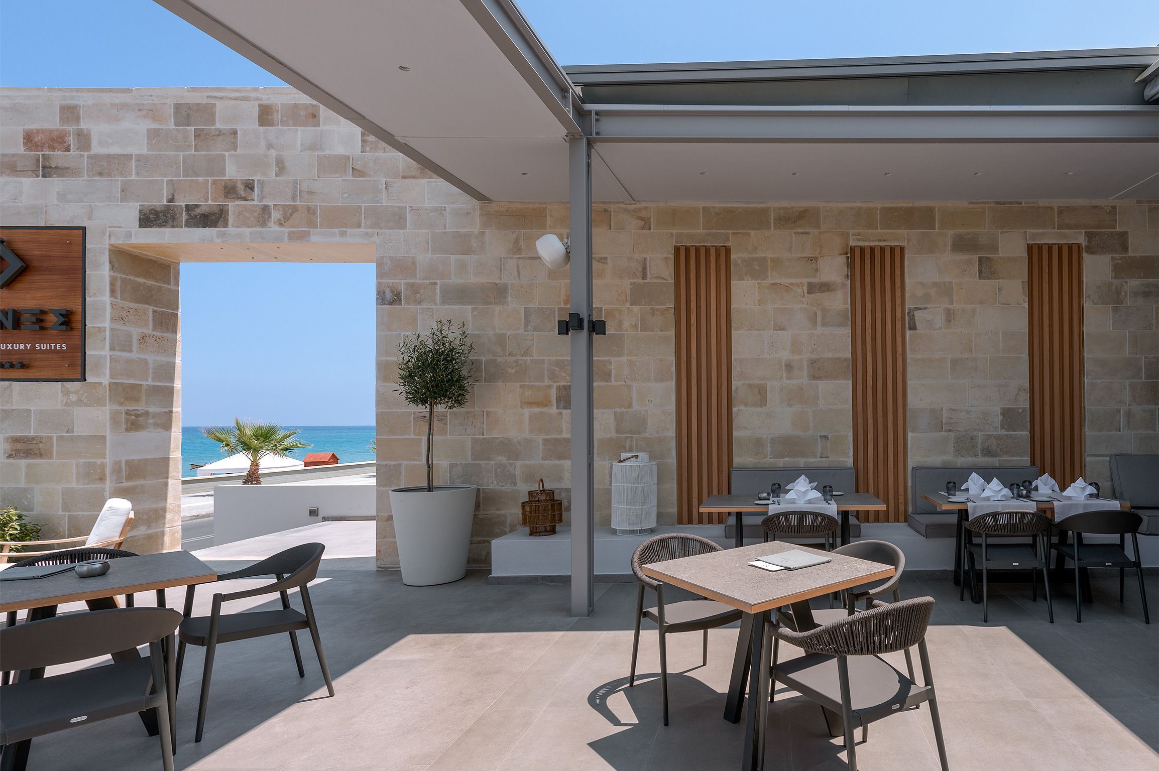 A modern outdoor dining area with stone walls, wooden accents, and a view of the sea.