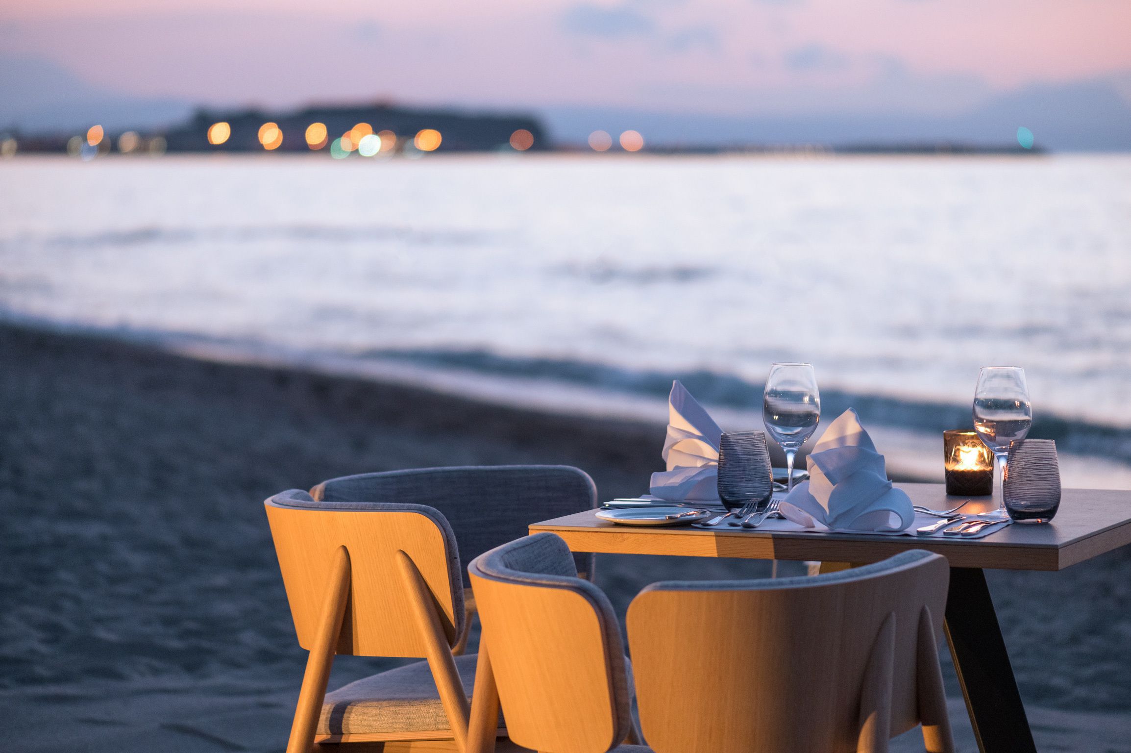 A table set for two with a candle, glasses, and a menu, overlooking a beach at sunset.