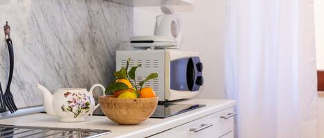 A kitchen with a white countertop, a microwave, a teapot, and a bowl of fruit.
