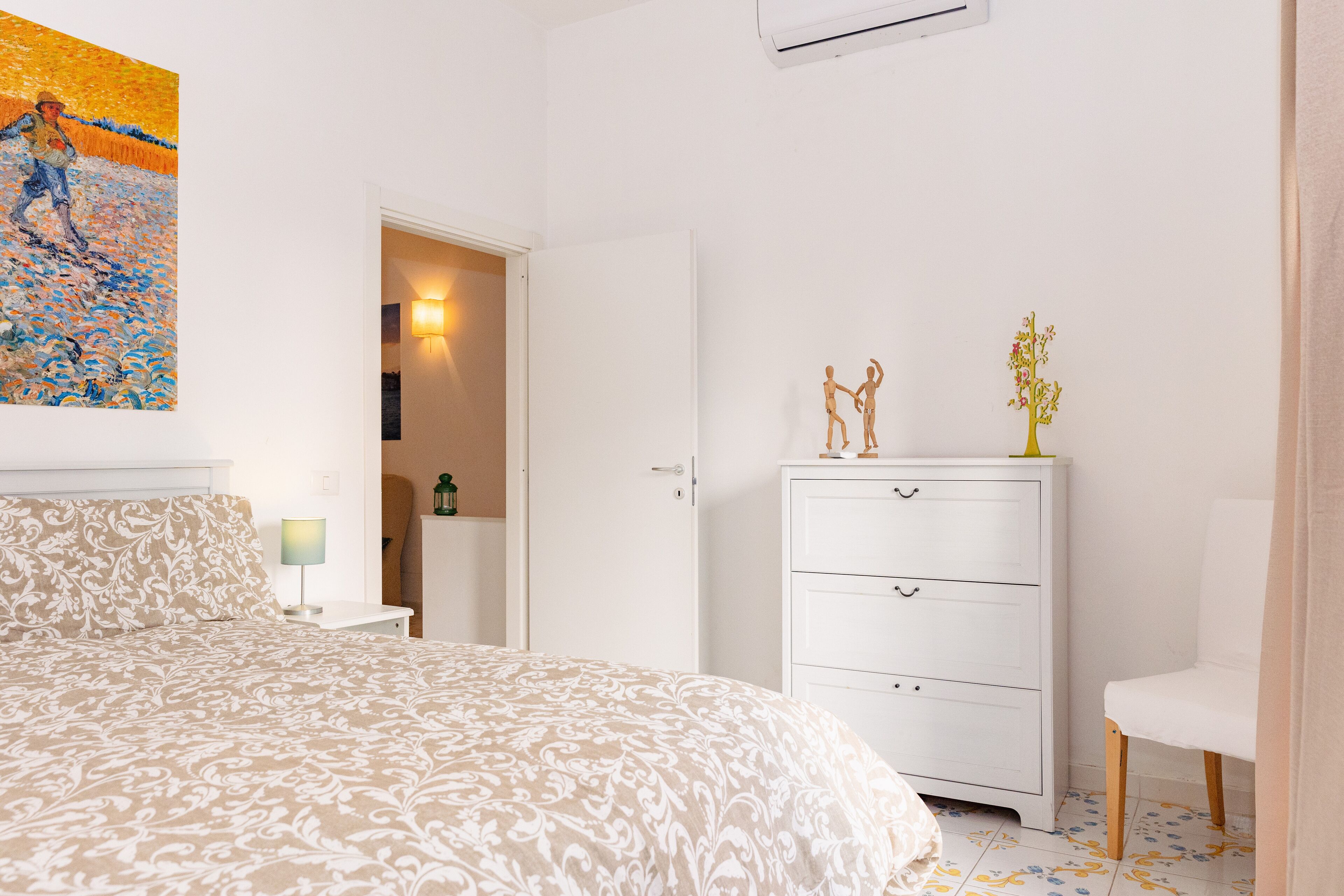 A bedroom with a patterned bedspread, a white dresser, a nightstand with a lamp, and a painting on the wall.